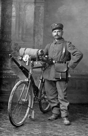 Aufnahme eines Arbeiters der Reutlinger Spinnereihülsen- und Spulenfabrik, der im September 1915 „als kriegsfreiwilliger Radfahrer eingerückt“ ist. Das Rad hat eine Karbidlampe, für das Gewehr ist eine Halterung am Rahmen angebracht, auf dem Lenker ist eine Decke festgezurrt. Im Hintergrund ist eine Foto-Tapete, das Rad und die Uniform zeigen deutliche Manöver- oder Gebrauchspuren.