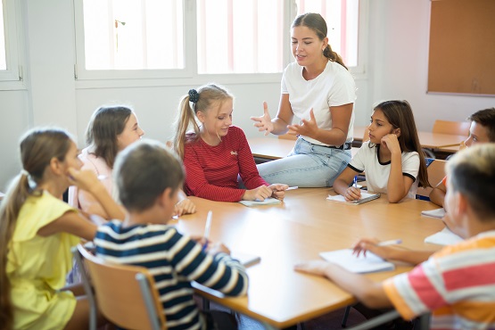 Young female teacher conducting lesson with interested preteen children sitting around common desk in classroom Symbolbild: Junge Lehrerin und Schüler